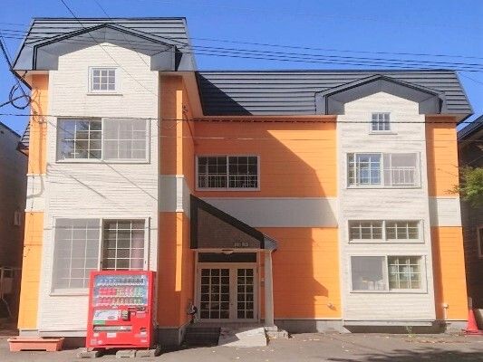 オレンジの2階建ての建物にはグレーの屋根が付き、赤い自動販売機も置かれている。玄関先には屋根が付き、スロープが設置されている。