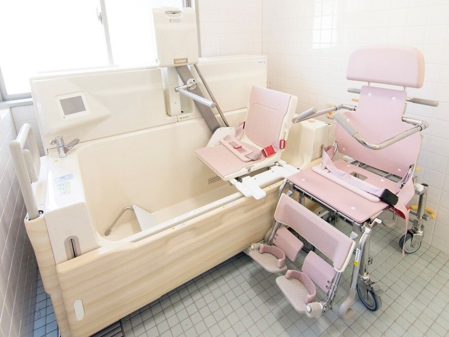 白とピンクの機械浴が設置されたタイル張りの浴室には窓が付き明るい。白い浴槽にはリフトが設置され、横には浴室用車いすがある。