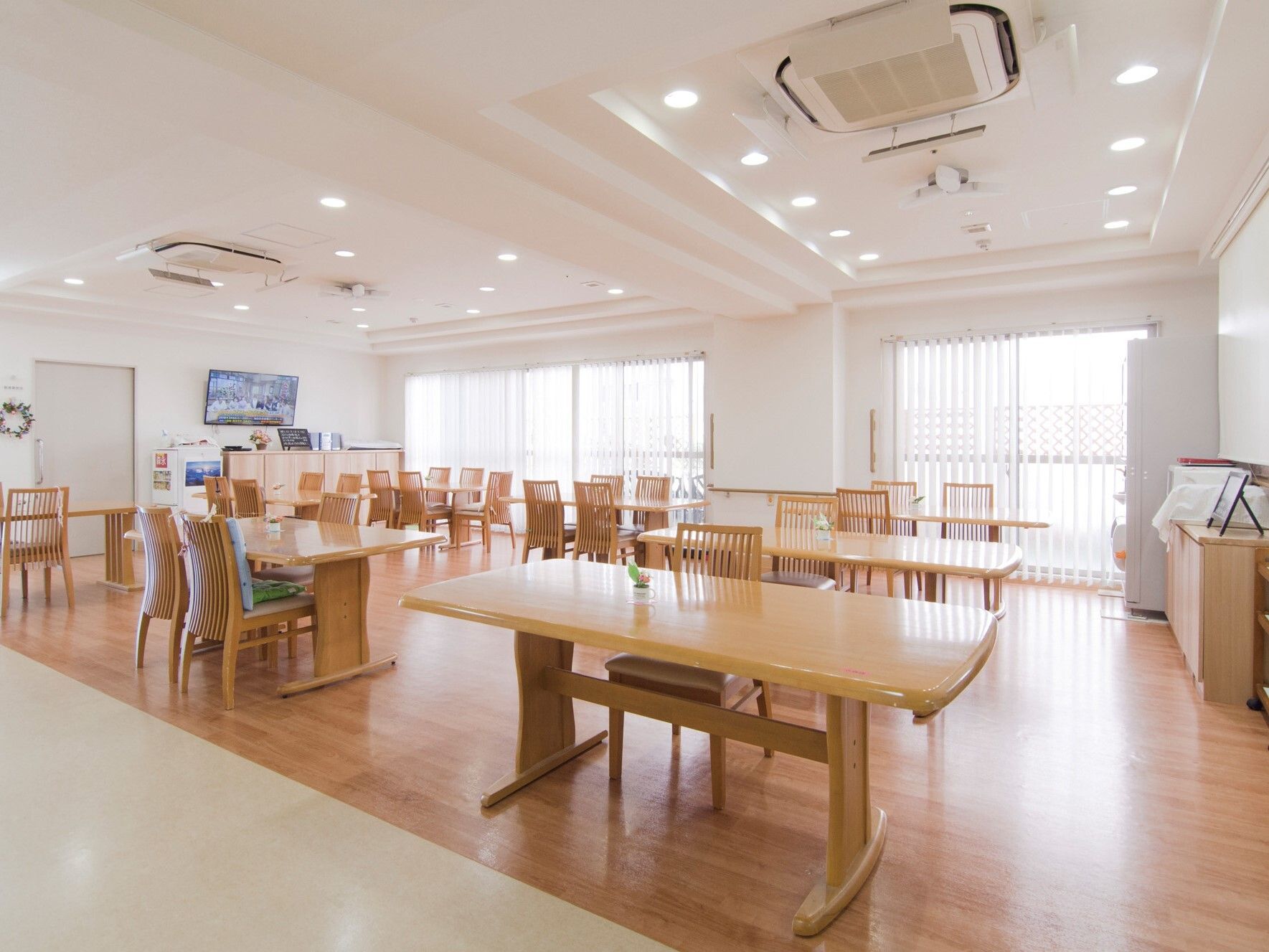 大きな窓が付いた明るい雰囲気の食堂。たくさんのダイニングテーブルが並び、壁には大きなテレビが付き、天井には業務用エアコンがある。