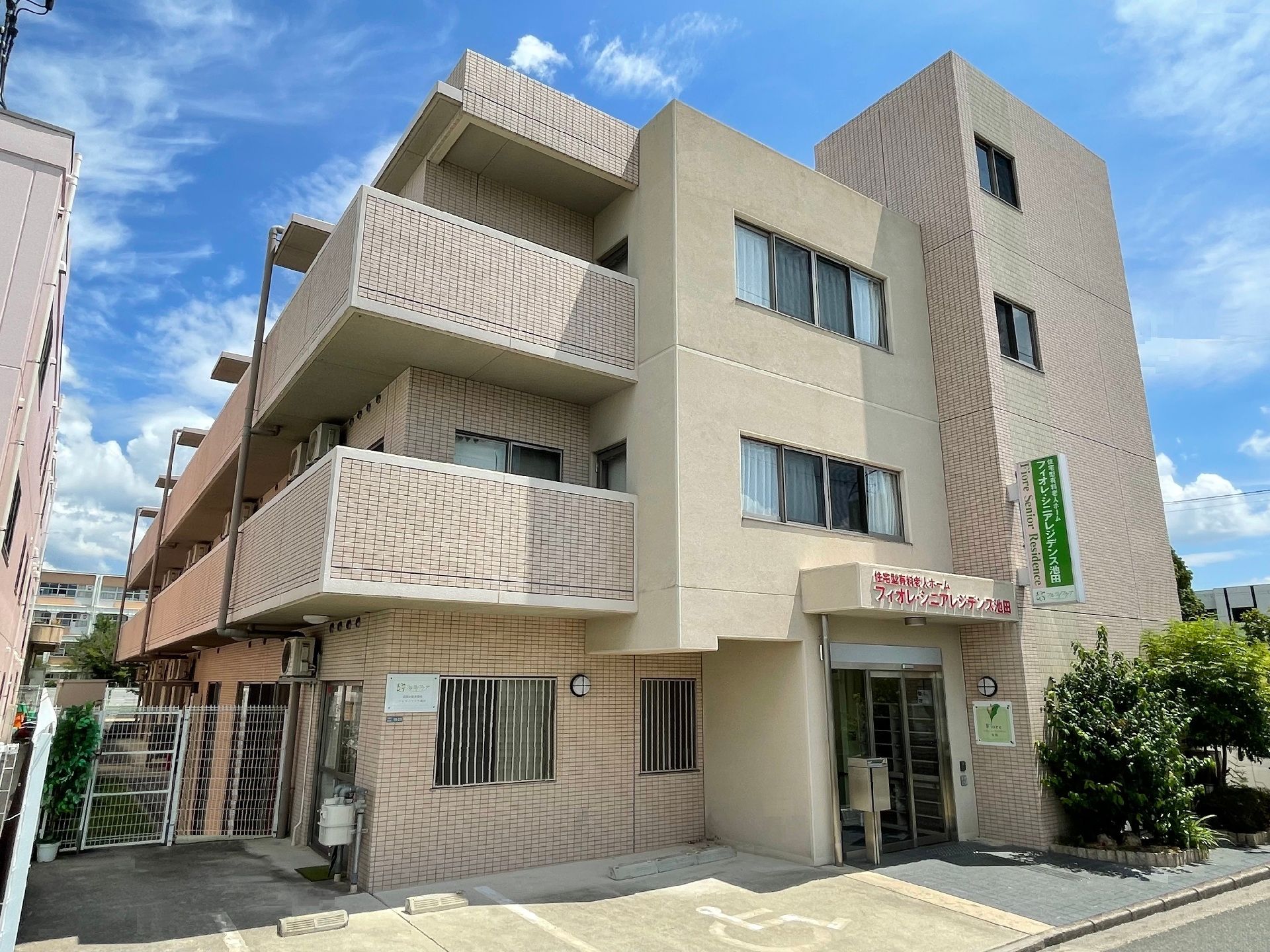 重厚感のある建物はベージュで玄関の屋根には赤い文字で施設名が書かれ、その横には緑が植えられている。各フロアにベランダが付いている。