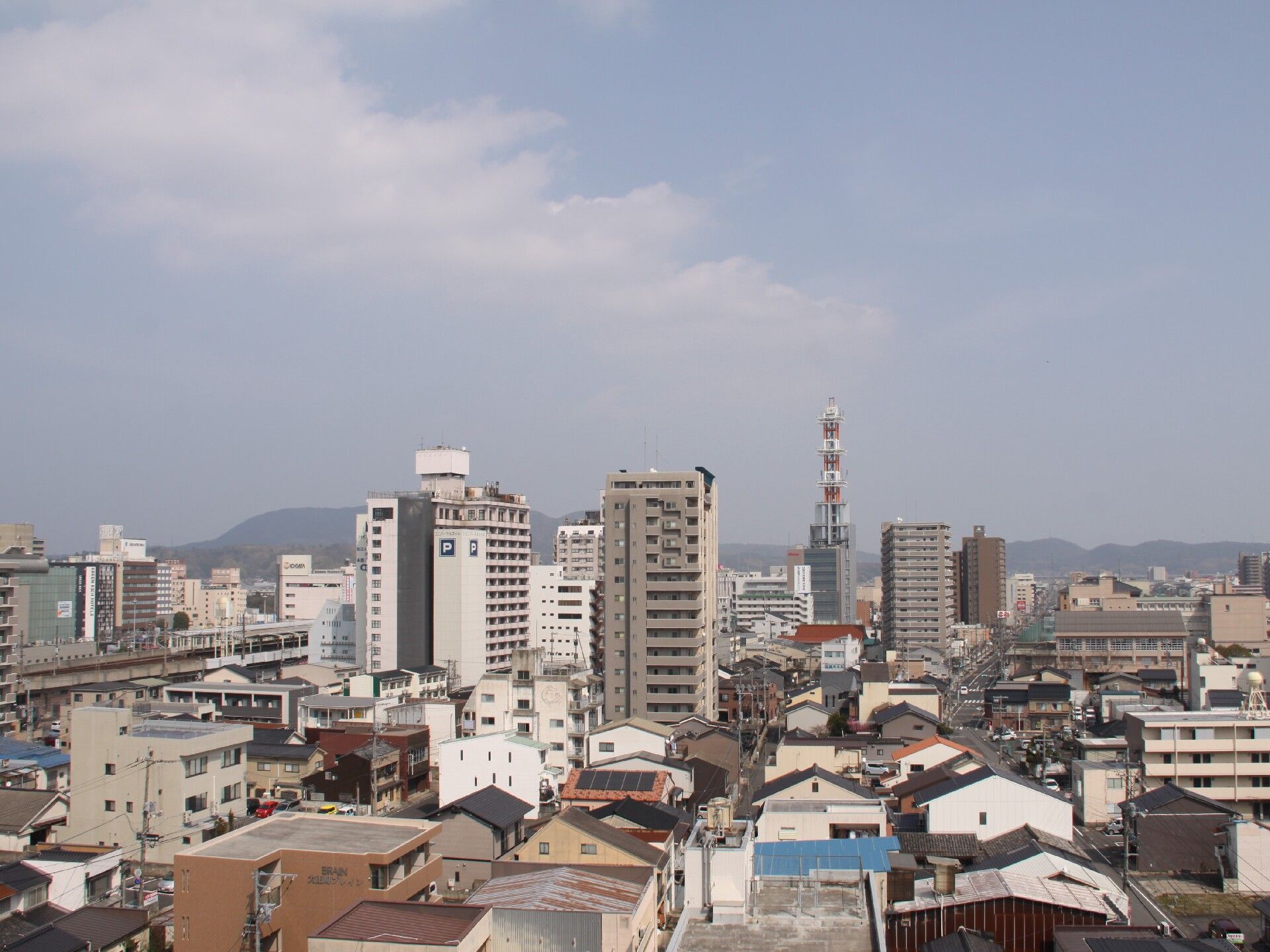 住宅が多くあるエリアの中央にビルなどが建ち並びぶぎやかな場所。奥には山々が連なっている。