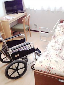 介護用ベッドには寝具がセットされ、近くには車いすが置かれ、奥にはデスクセットが置かれている。