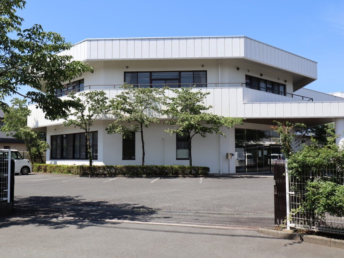 白い建物は2階建てで敷地内はアスファルトで整備され、門も付いている。建物周辺には緑が植えられ、駐車スペースもあり、2階部分にはテラスもある。