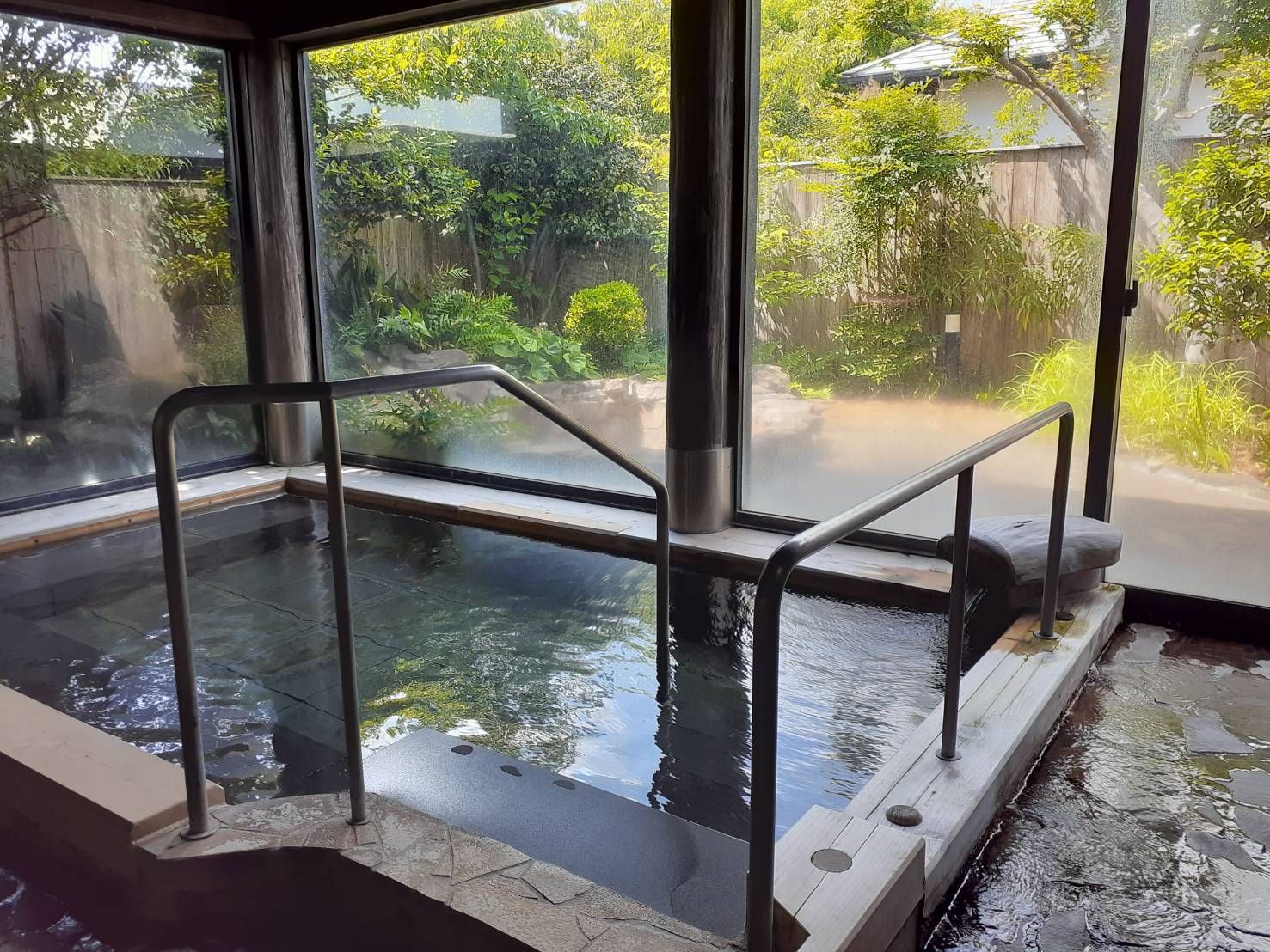 大きな湯船には手すりが設置され、大きな窓の外に見える和風庭園や石畳のフロアが和風の旅館のような雰囲気。壁には桶もかけられている。