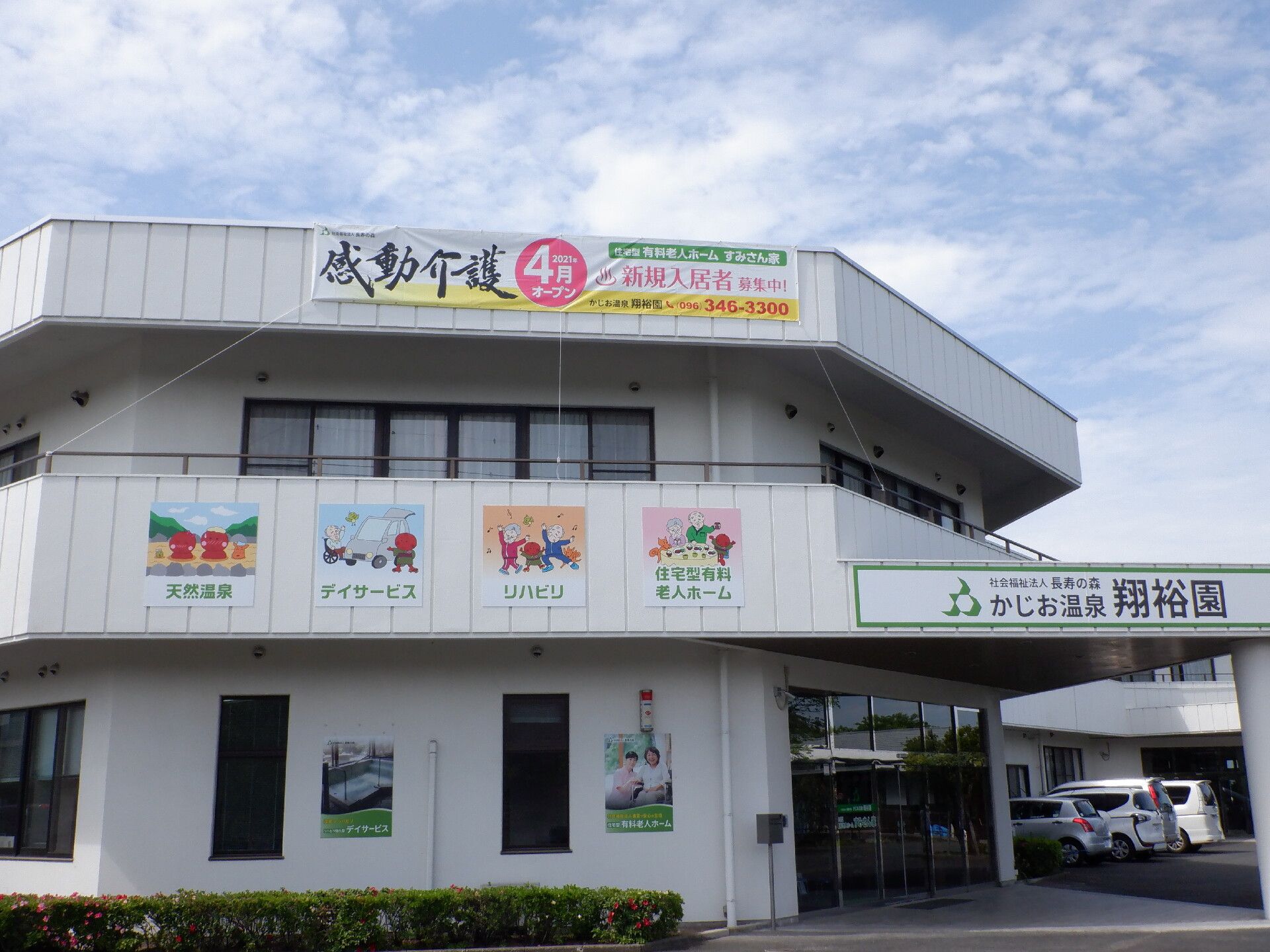 白い建物は2階建てで木がたくさん植えられた敷地内の駐車スペースには白い車も止まっている。玄関には自動ドアが設置され、屋根も付いている。