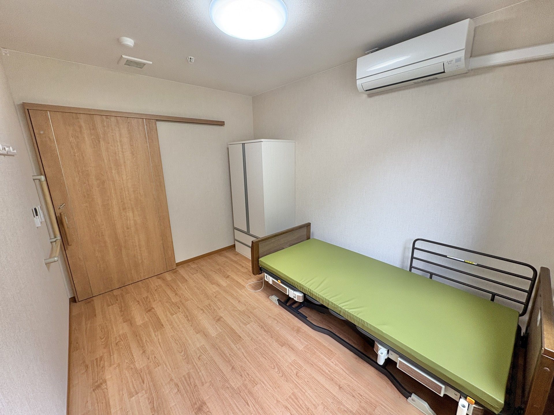 日当たりのいい屋内で車いすの高齢女性が誰かに向かって笑顔で手を振っている。そばにいる若い女性スタッフも笑顔で同じ方向を見ている。。