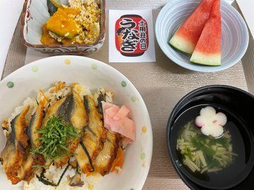 大きくカットされたウナギが乗っているうな丼に吸い物、カボチャの煮物、スイカなどが並んでいる。