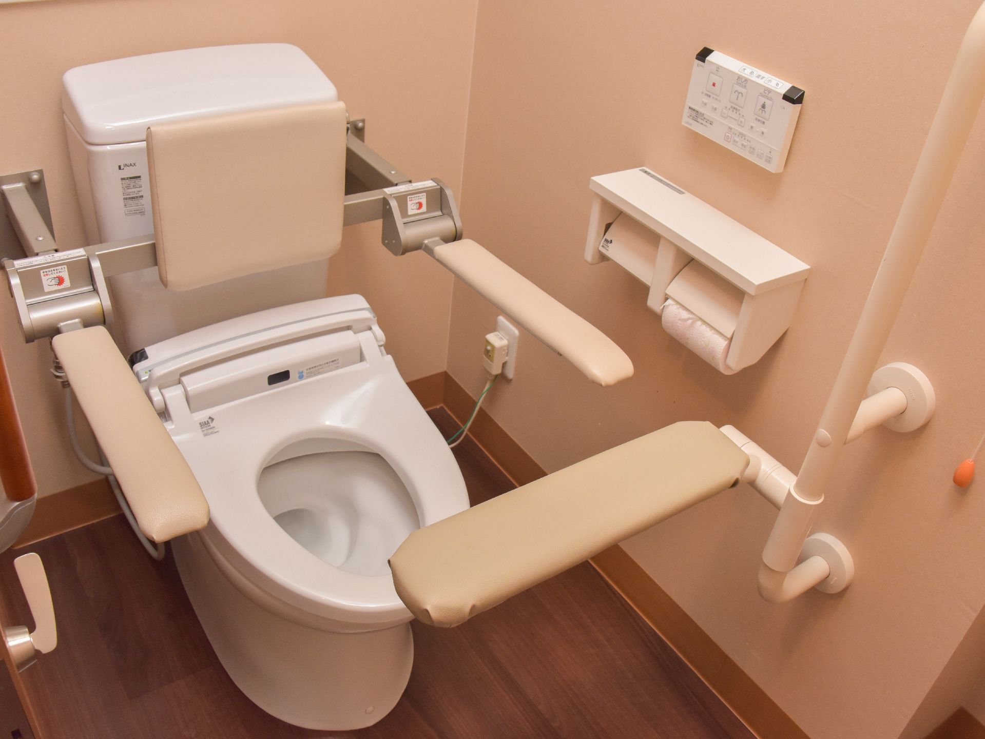 個室の洋式トイレで、手すりと背あてがついているため立ち居がらくである。壁面にウォシュレットの操作パネルが取り付けてある。