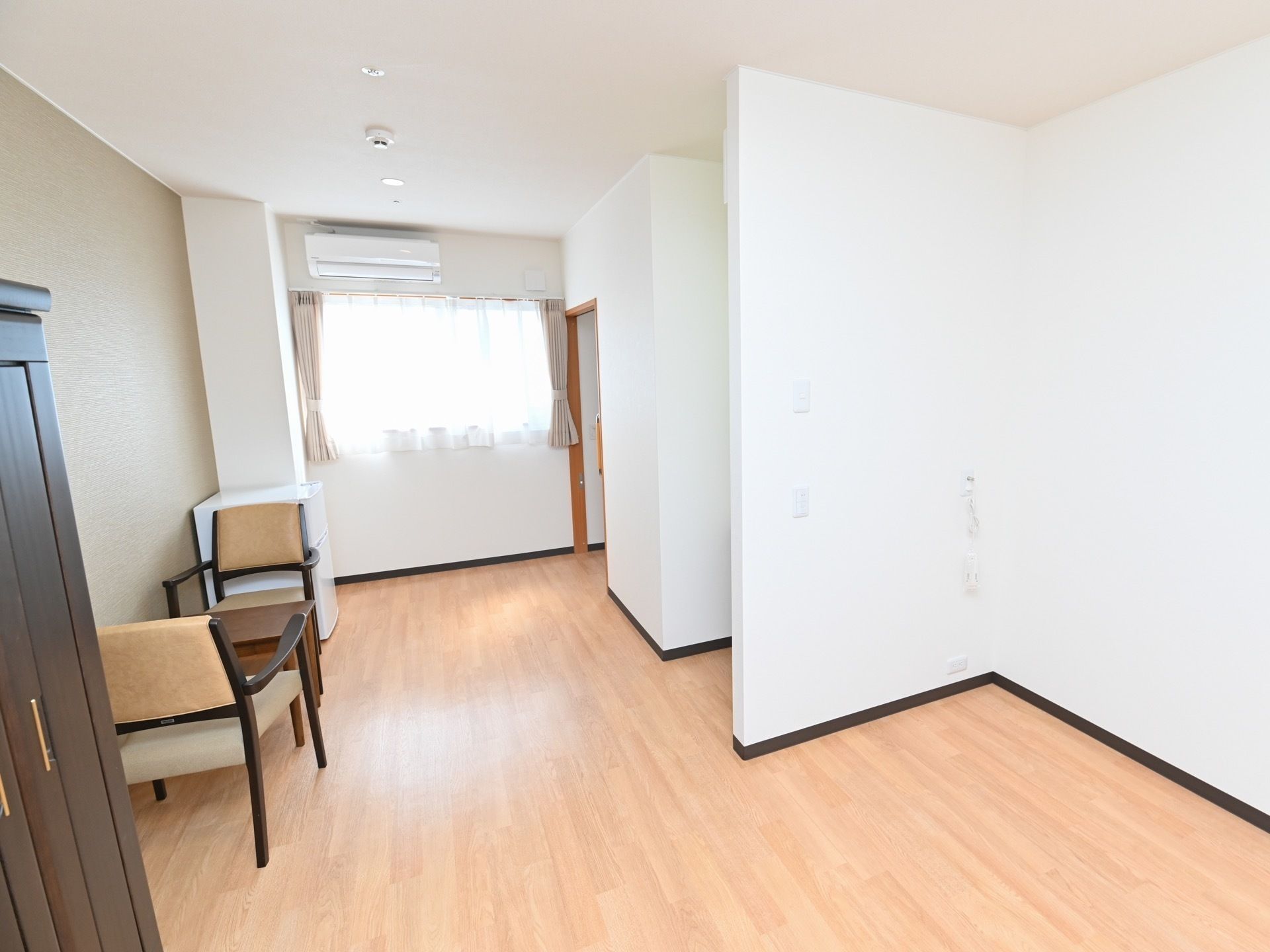 窓から光が差し込む明るい部屋である。部屋にはテーブルや椅子、エアコンや収納棚などが取りつけられている。