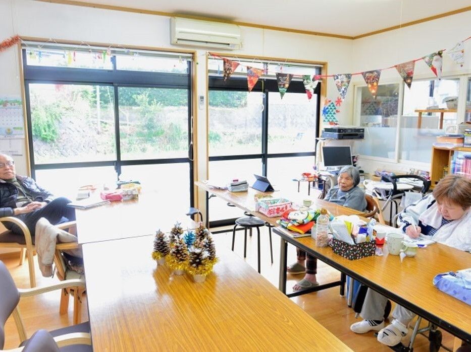 男女の入居者が数名共有スペースでくつろいで座っている様子。手作りのガーランドや入居者の作品が飾られている。