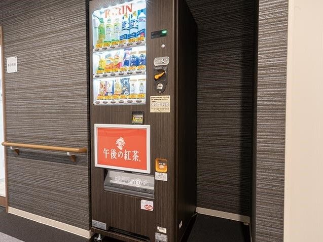 壁と同じダークグレーの自動販売機が置かれていて「午後の紅茶」と書かれイラストが描かれた赤い大きなステッカーが貼られている。