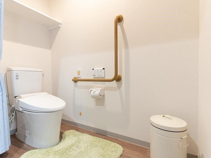 木製のエル字型の手すりやオレンジの呼び出しボタンが付いた広いトイレにはマットが敷かれ、ゴミ箱もある。