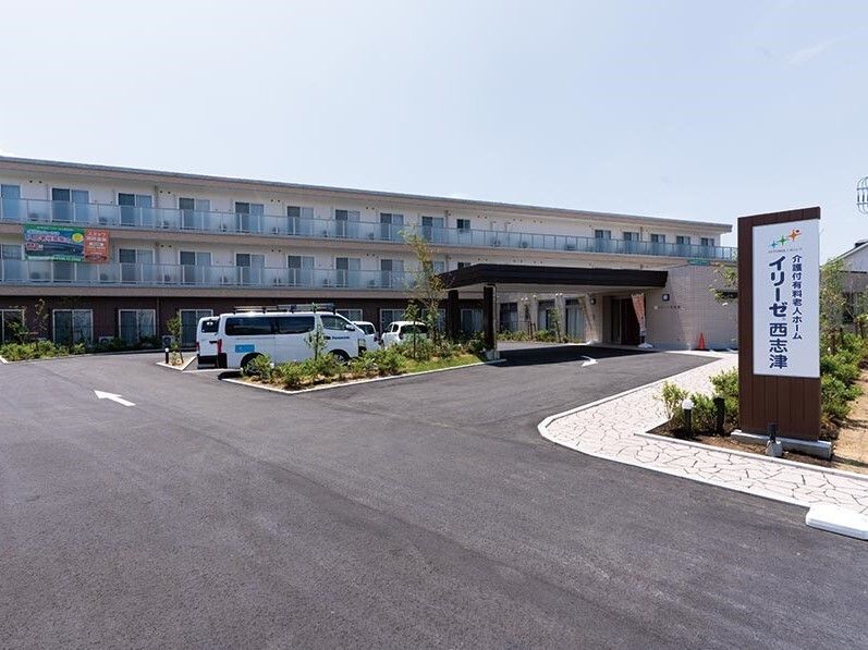 広い駐車場が付いた3階建ての建物は横に長く、敷地内にはたくさんの草木も植えられている。白い看板も建っている。