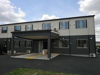 2階建ての建物はフロアにより外壁の色が異なり、広い敷地内には駐車場があり、玄関には大きな屋根がある。