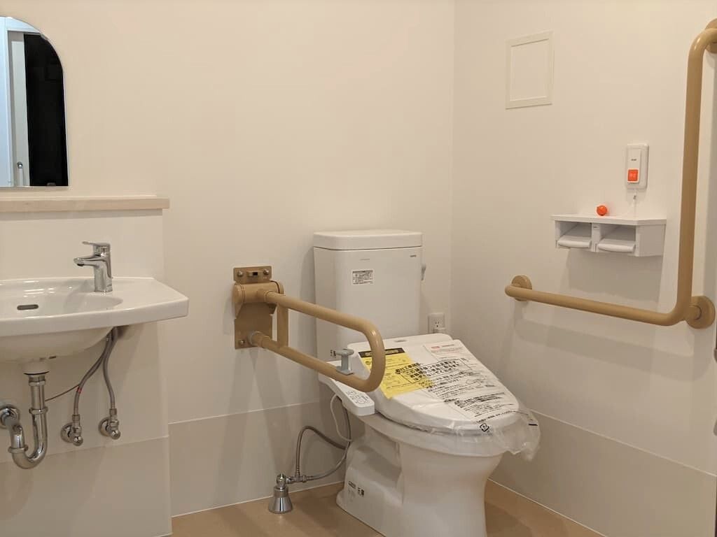 鏡付きの手洗い場が付いた広いトイレには両サイドに手すりが設置されたトイレがあり、呼び出しボタンやトイレットペーパーホルダーも付いている。
