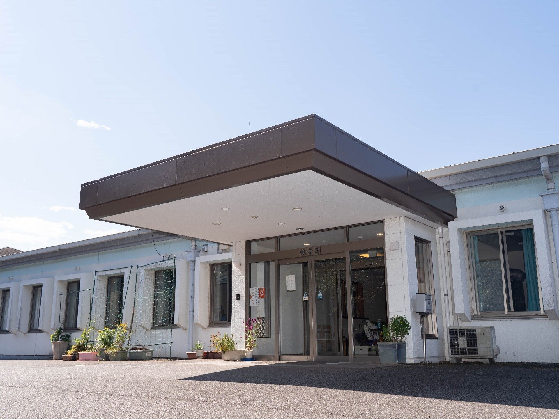 2階建てのベージュ系の建物。1階部分にはウッドデッキのテラスがあり、テーブルセットが置かれている。