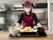 エンジのキャップとエプロンを身につけた女性スタッフが食事が並べられたトレーを差し出している。天ぷらなどがある。