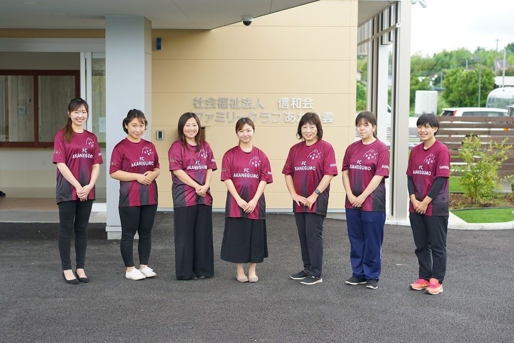 エンジのそろいのTシャツを着た女性スタッフ立ちが横一列に並び、微笑んでいる。後ろには住宅の玄関がある。