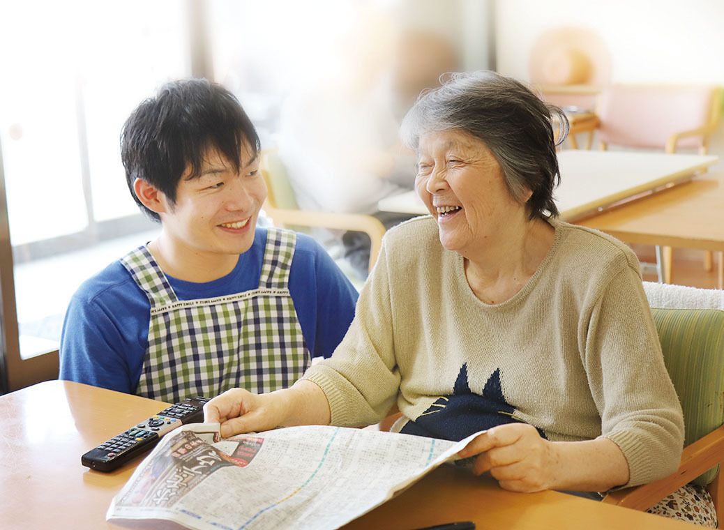 チェックのエプロンを付けた若い男性スタッフとテーブルに新聞を広げている高齢女性が笑顔で話している。