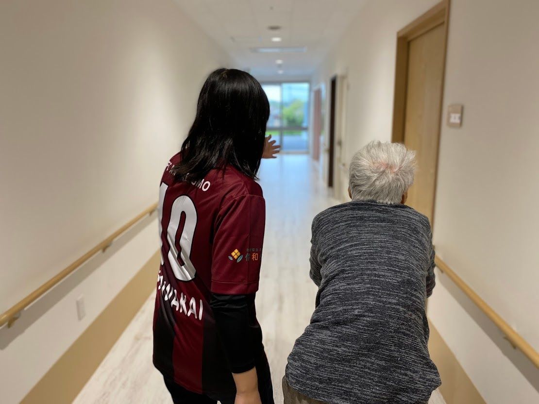 共有スペースのテーブルの上に新聞紙を広げている高齢女性と腰を下ろして目線を合わせている男性スタッフ。笑顔で話している。