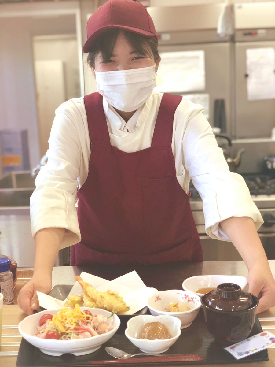 炊き込みご飯や天ぷらの盛り合わせなどが盛られた黒いトレーを差し出している厨房スタッフの女性は、エプロンとキャップを身につけている。