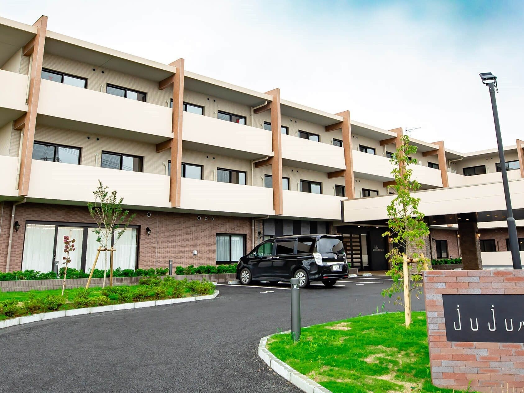 3階建ての建物はベージュとブラウンで各フロアにベランダがあり、駐車場も設置され、緑も植えられている。