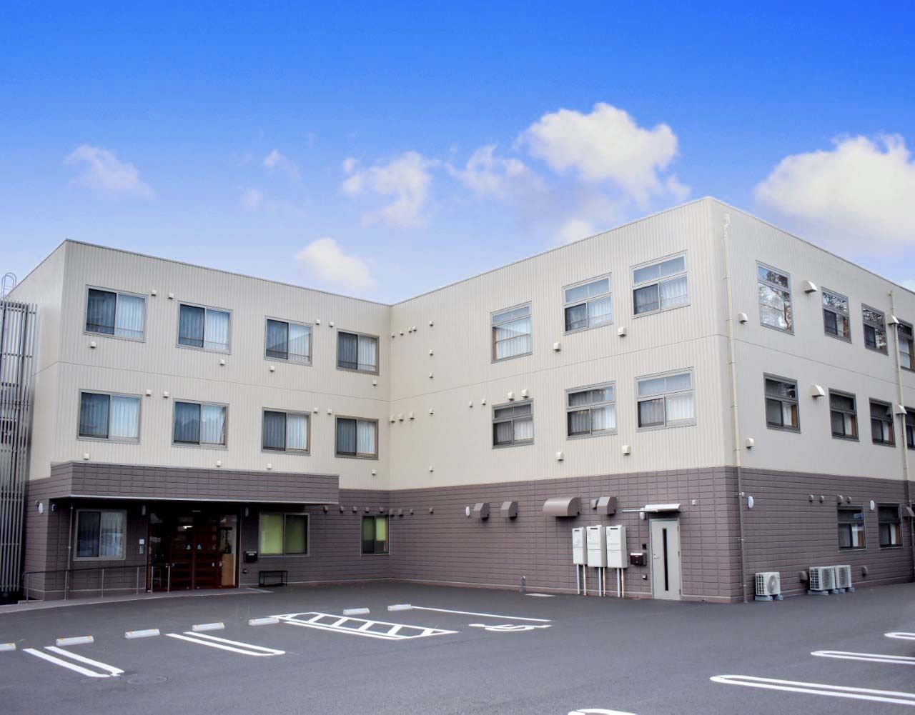 大きな駐車場が付いたエル字型の3階建ての建物はベージュ系。敷地内には駐輪場もあり、緑も植えられている。