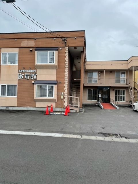 ブラウン系の2階建ての建物。外壁には白い文字で住宅名が書かれ、玄関にはスロープがあり、道路に面している。