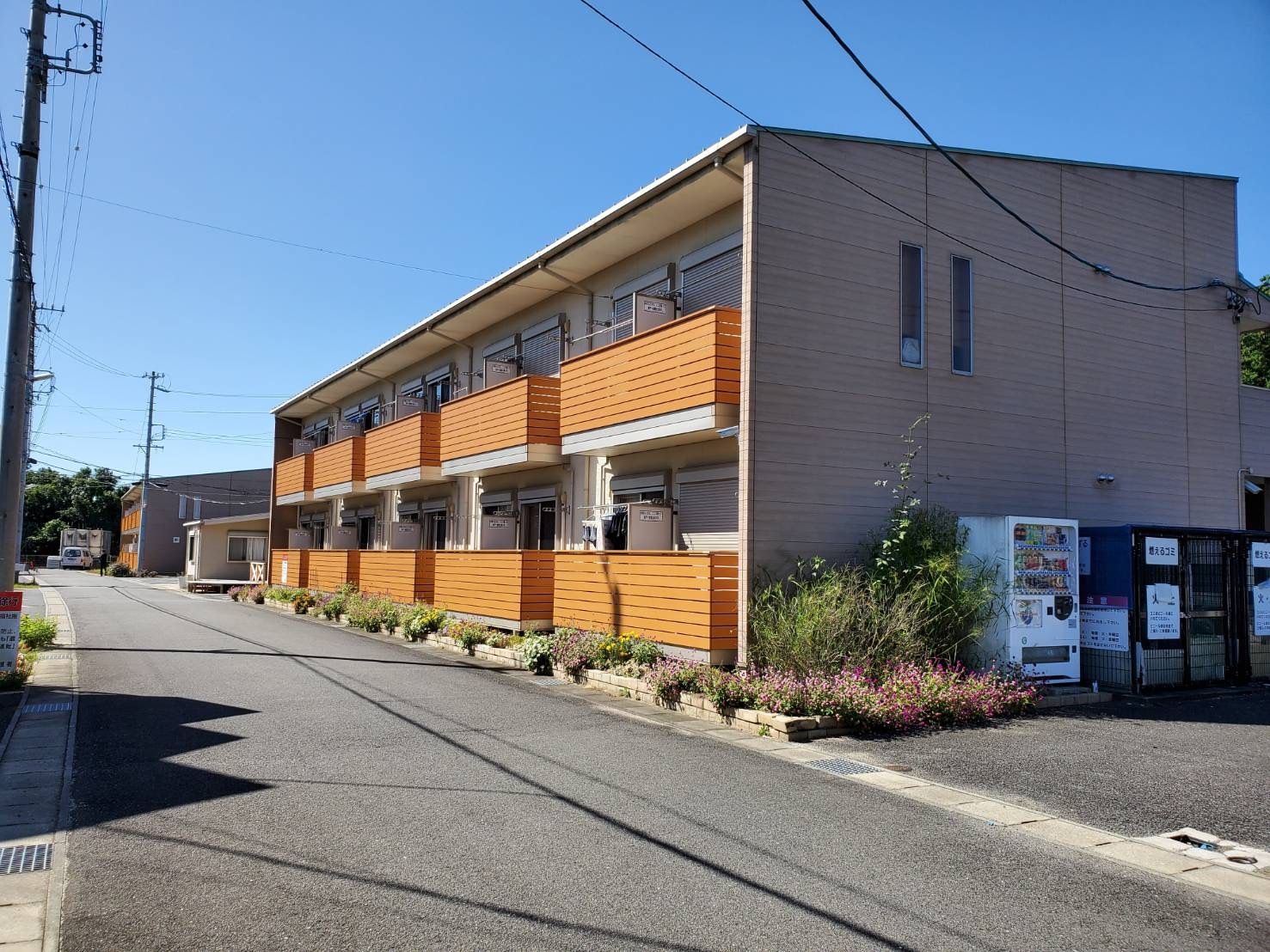 オレンジのバルコニーがそれぞれの部屋に付いた2階建ての建物。道路に面していて、敷地内にある駐車場の一角には自動販売機もある。