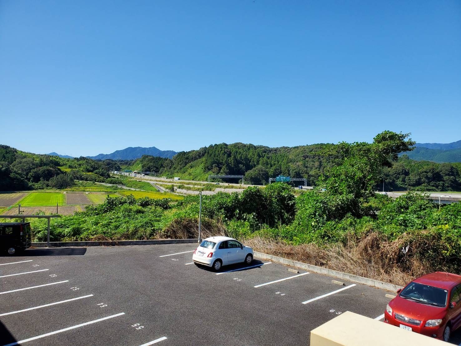 青い空の下に広がる緑豊かな風景。奥には山々が連なっていて、敷地内には白線が引かれた広い駐車スペースがあり、車が止められている。