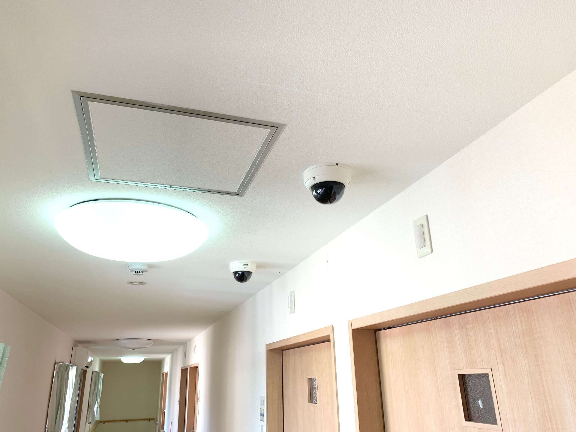 360度見ることができる黒い防犯カメラが２つ設置された廊下の天井。居室のドアは木製で、小さな小窓が付いている。壁は白く清潔感がある。