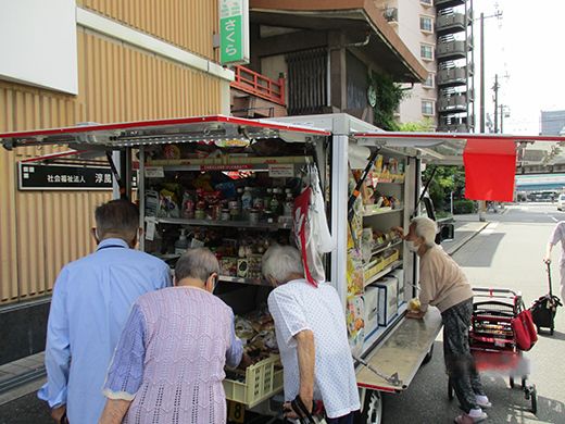 移動スーパーの車が止まり、高齢者たちが真剣に品定めをしている様子。中にはシルバーカーを使用している人もいる。
