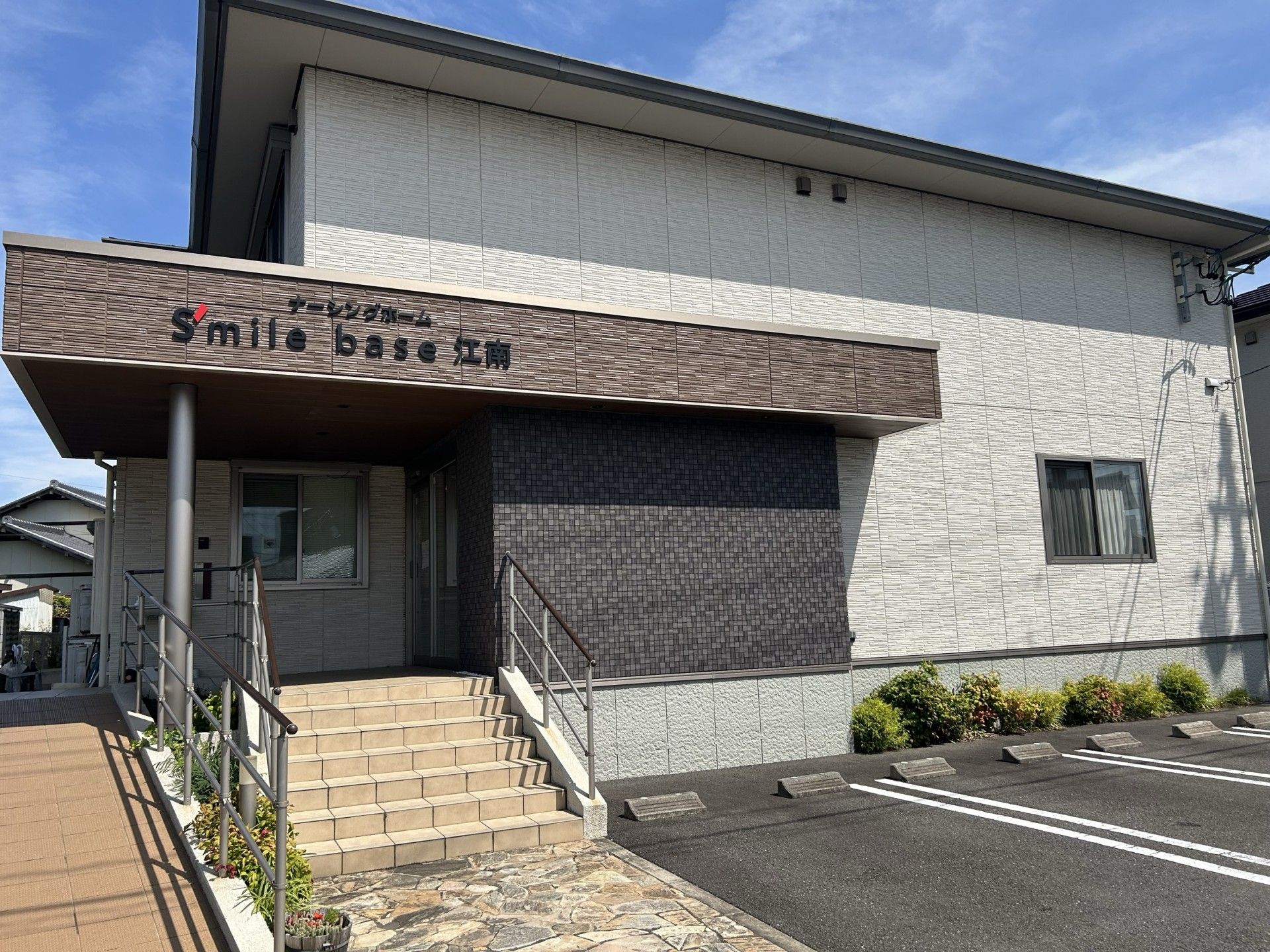 白い外壁にレンガの模様が描かれたブラウン系の屋根が付き、ホーム名も書かれている。玄関には手すり付きのスロープと階段が付いている。