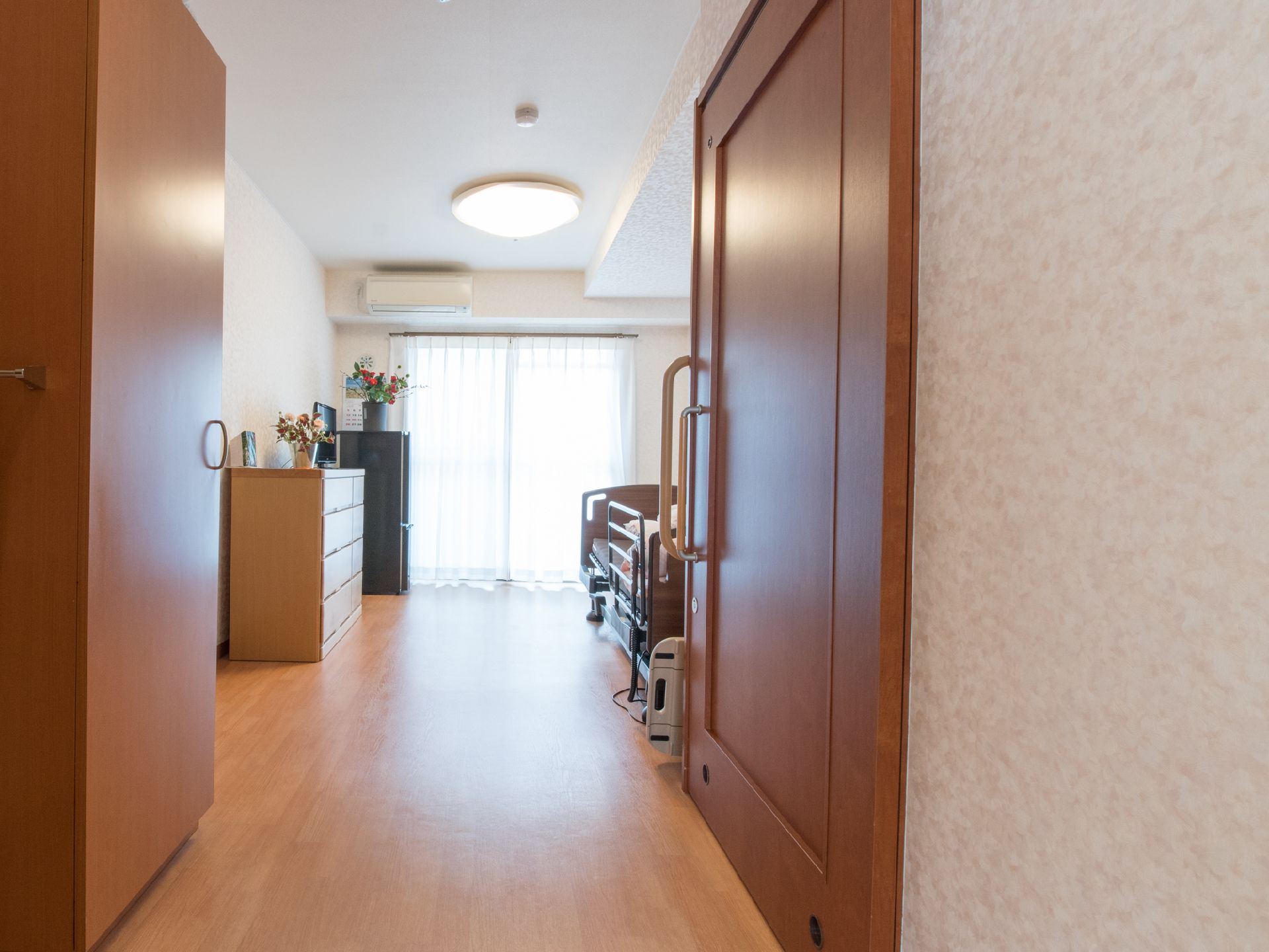 茶色と白基調の内装である。薄手のカーテンから明るい光がさしこんでいる。窓の左上にはエアコンが設置されている。家具の上には花が飾られている。