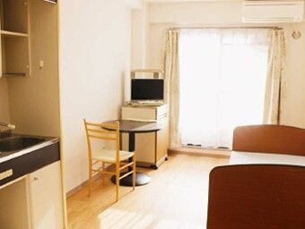 大きな窓が付いた明るい雰囲気のフローリングの部屋。車いす対応のキッチンや寝具付きのベッド、テーブルセットやエアコンがある。