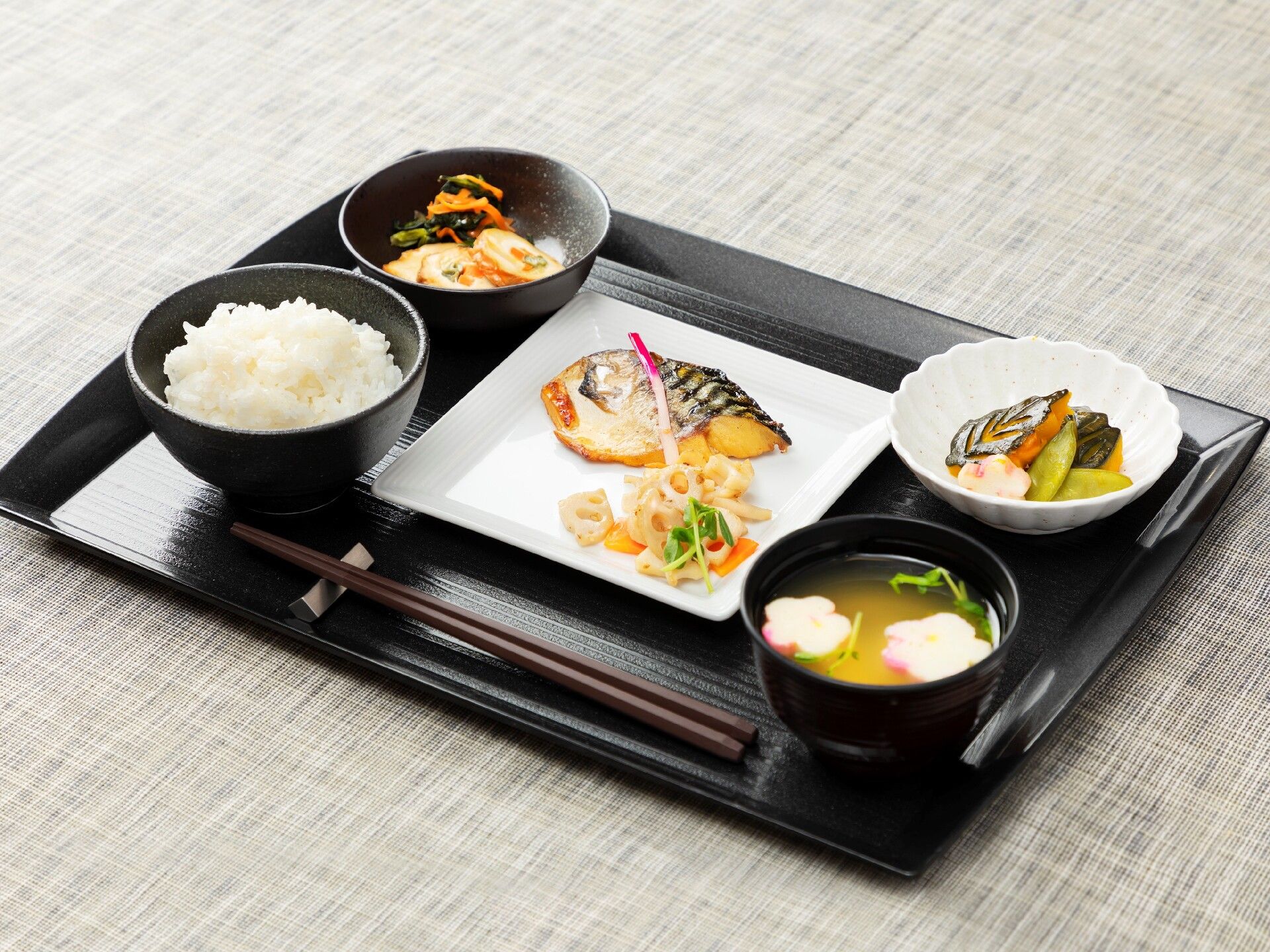 白いご飯に味噌汁、サバの塩焼き、野菜の和え物が数種類がそれぞれ皿に盛られ、黒いトレーに置かれている。