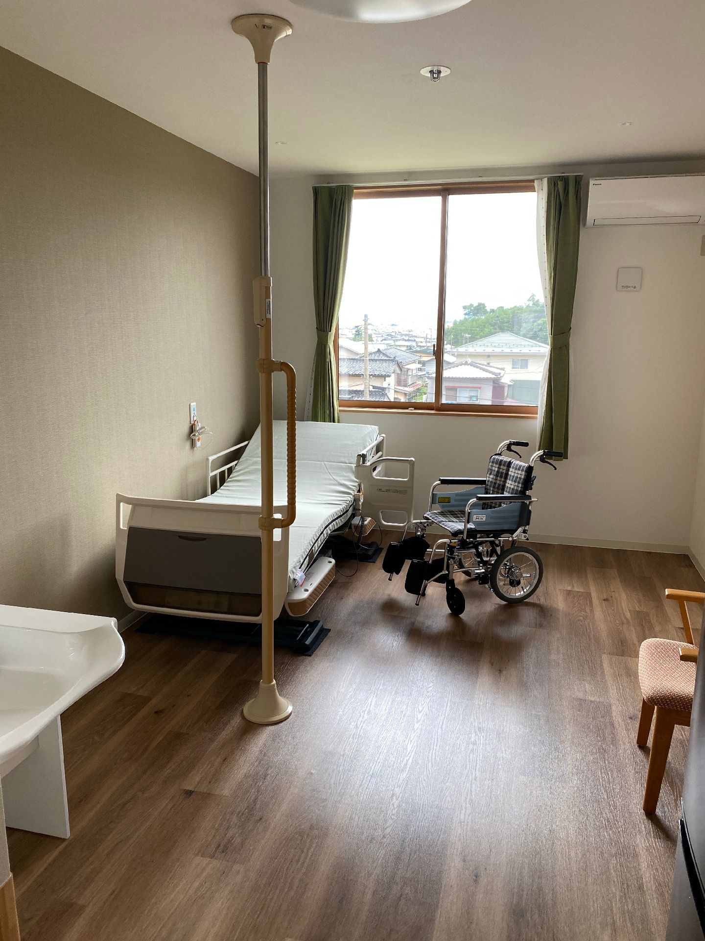 カーテンが付いた大きな窓が設置されているフローリングの部屋には介護ベッドやいす、車いす、エアコンなどがある。