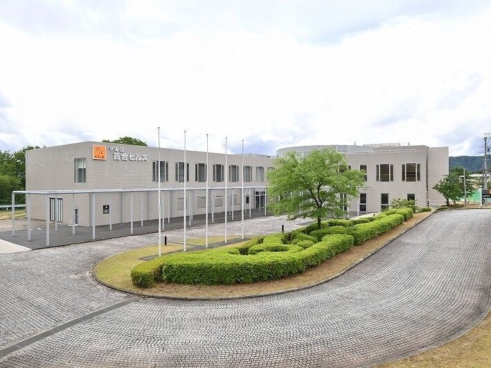 グレーの2階建ての建物は緑に囲まれていて、建物の周りには減洲があり、緑地スペースを中央に置いたロータリーもある。