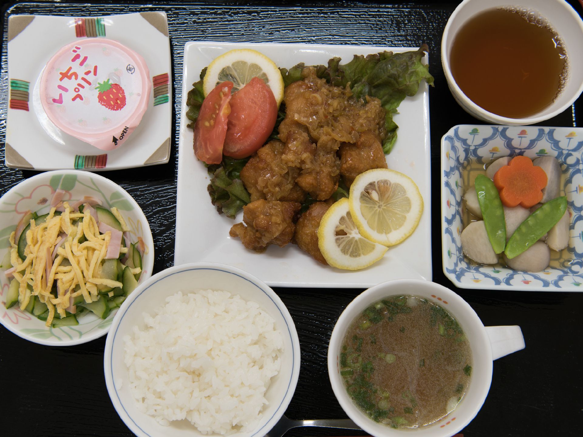 食事の写真。主菜はネギソースのから揚げ。副菜はさといもの煮物とハム・きゅうり・玉子の和え物。いちごプリンも付いている。