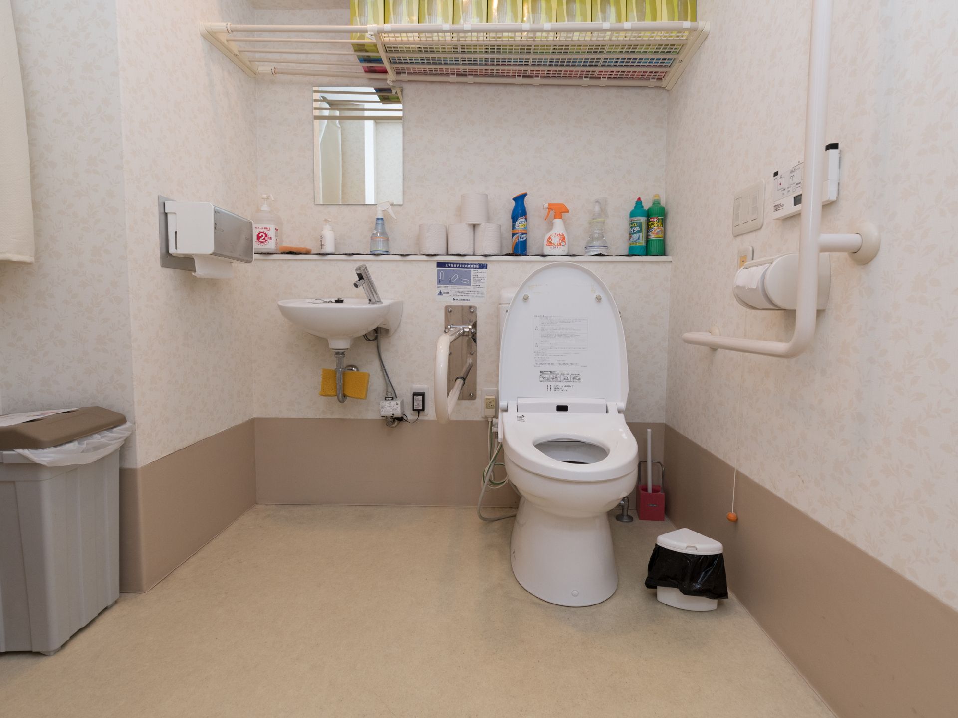 広くてゆったりとしたトイレ。棚には洗剤やトイレットペーパーが置かれている。壁には手すりが設置されている。