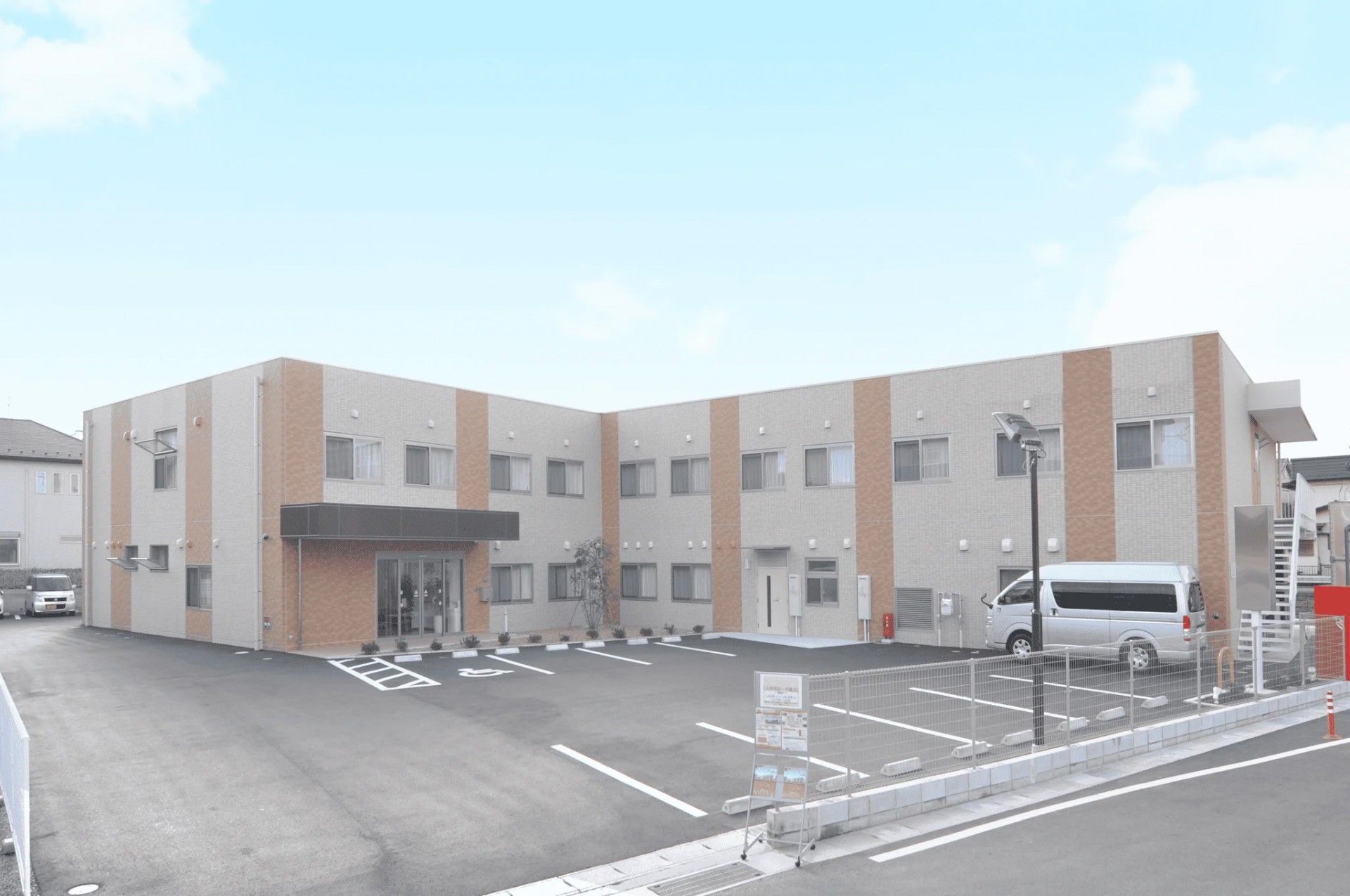 駐車場がある2階建ての建物はエル字型でグレーの外壁にブラウンのストライプが入っている。