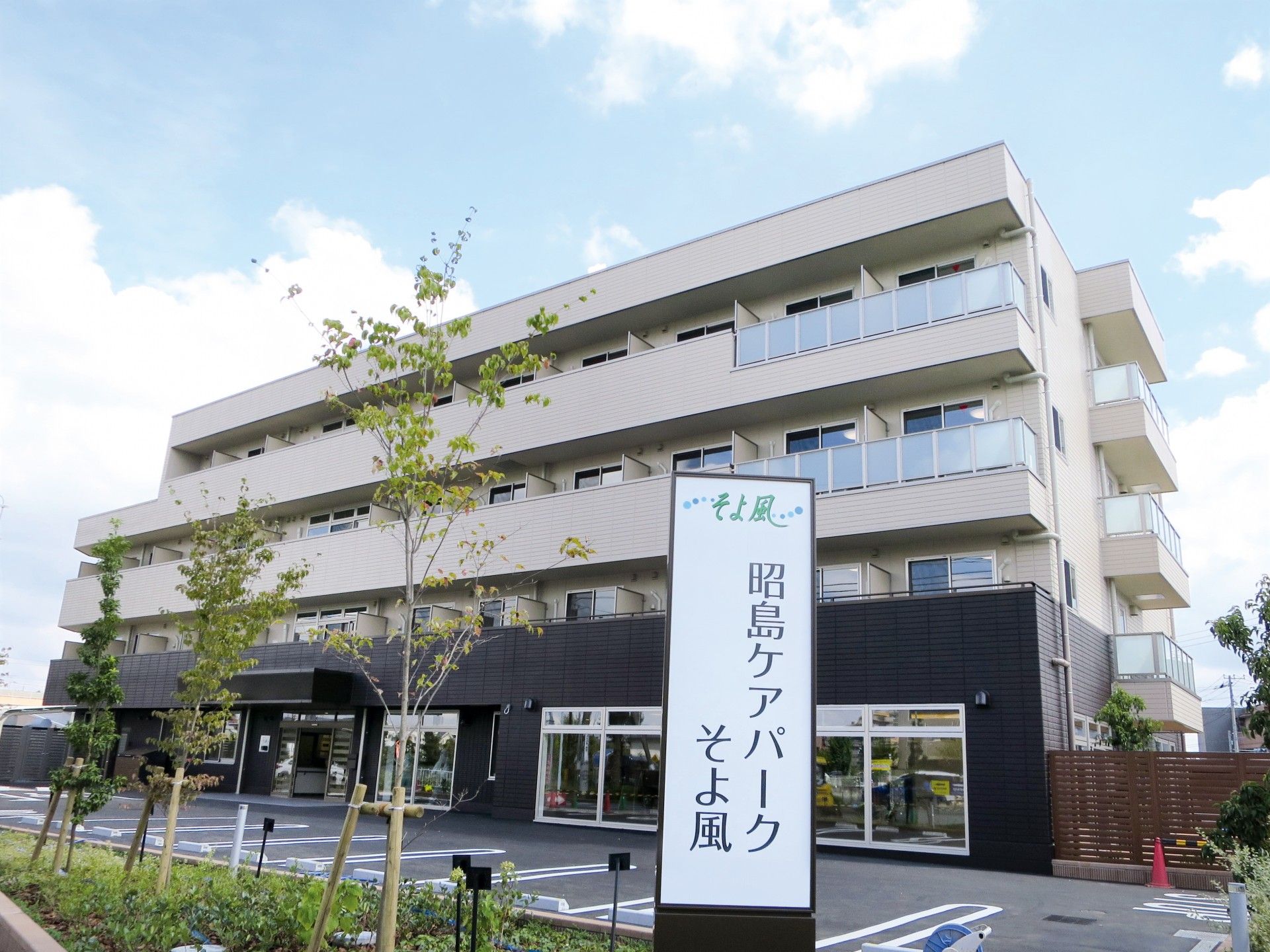 道路に面した白とブラウンの4階建ての建物。建物の前には大きな看板があり、周辺にはたくさんの木が植えられている。