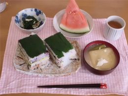 押し寿司やみそ汁、スイカのスライスや野菜の和え物などがピンクと白のランチョンマットの上に並べられている。