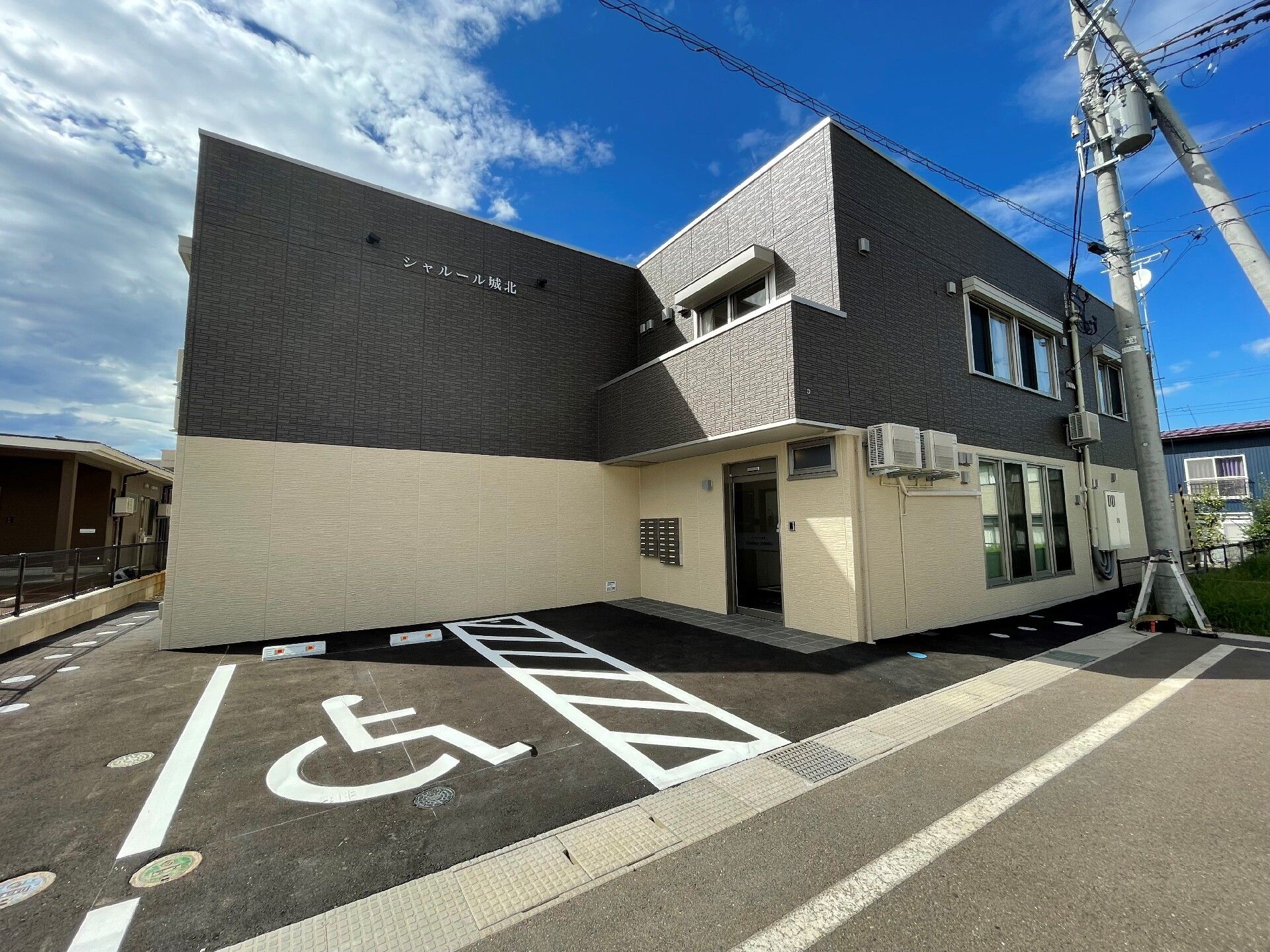 ブラウン系の建物は2階建てで、広い駐車場が付いている。玄関には屋根があり、玄関の前の駐車スペースには車いすのマークがある。
