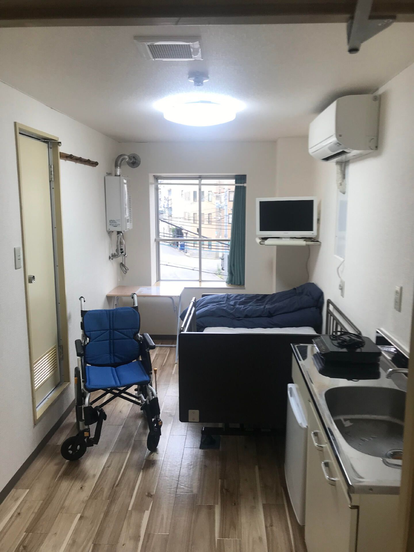 車いすがセットされたフローリングの部屋には寝具付きのベッドやテレビ、キッチン、エアコンなども付いている。