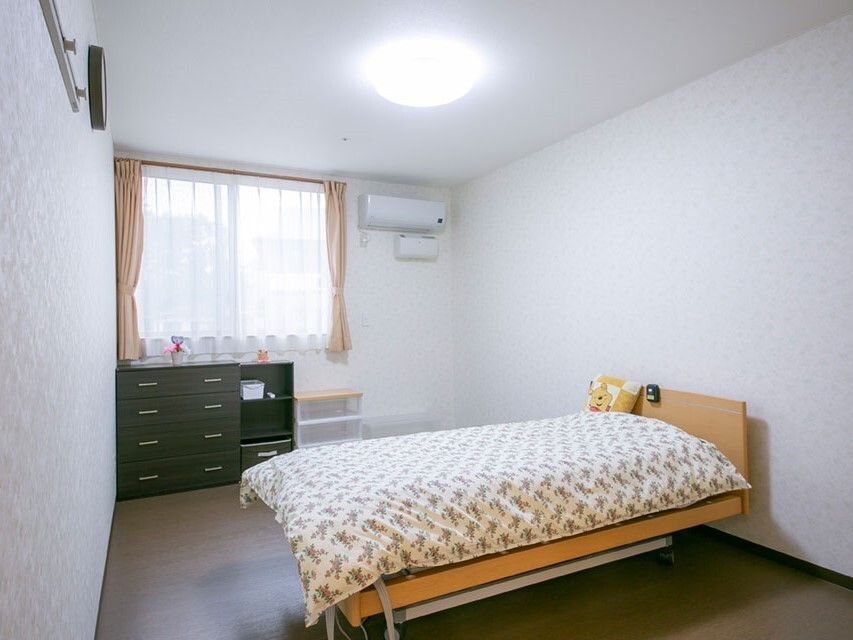 エアコンが付いた部屋に寝具付きのベッドが置かれ、タンスも設置されている。レースカーテンとカーテンが付いた窓もある。