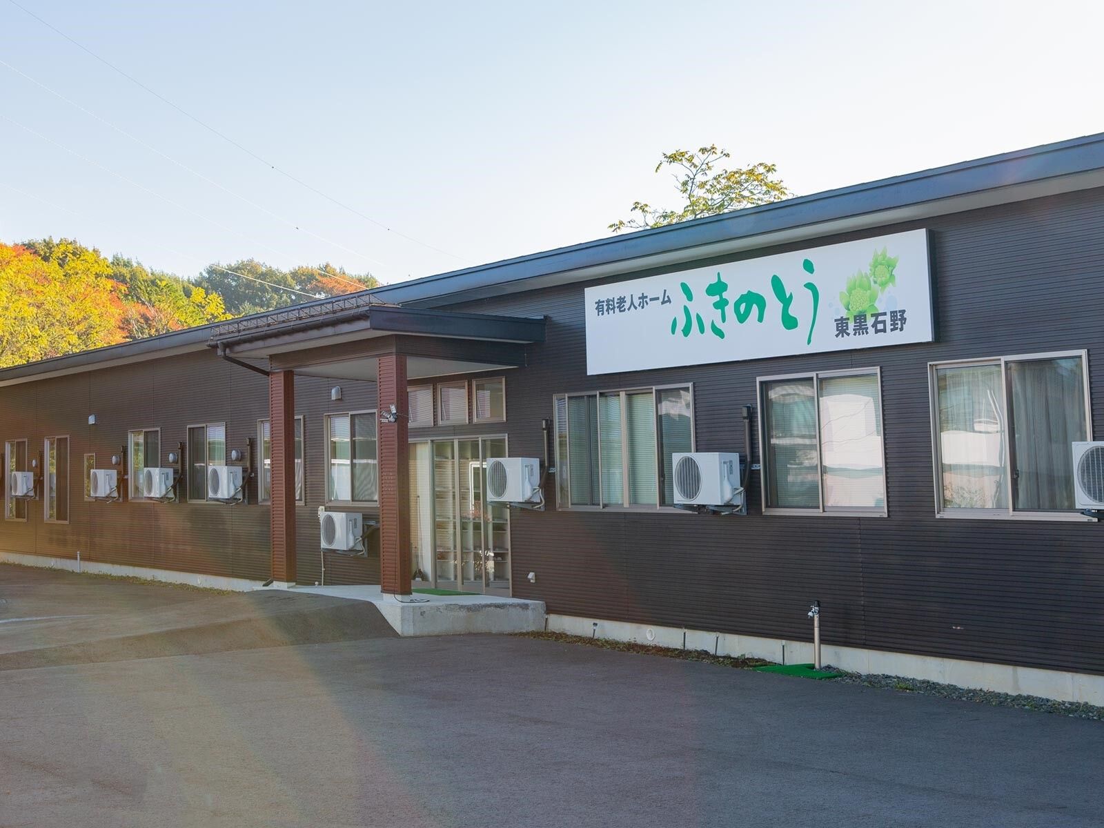 平屋建ての建物の周りには緑が多くあり、紅葉した山が後ろにある。建物にはホーム名が書かれた看板も設置されている。