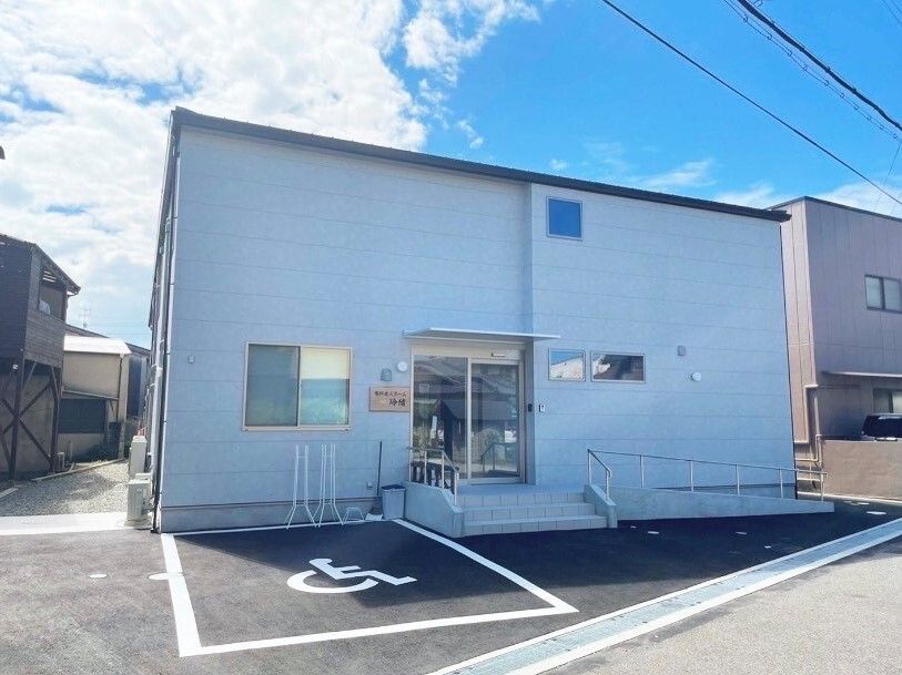 日当たりのいい場所に建っているライトブルーの外壁の建物の玄関には手すり付きのスロープと階段が付き、駐車場がある。