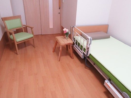 マット付きの介護ベッドが置かれたフローリングの部屋にはいすがあり、花が飾られた小さなテーブルもある。