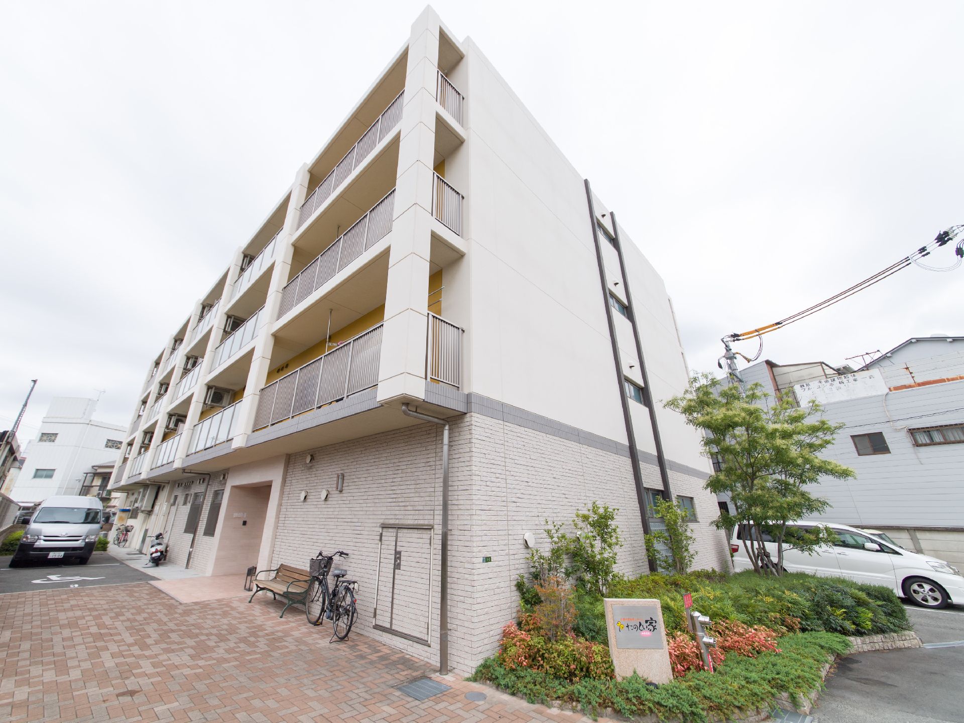 建物は、4階建てのRC造である。エントランスの近くにベンチが置かれている。施設名を表示した看板が、植栽の中にある。